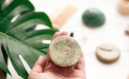 Zero waste. Choice plastic free eco products. Hand holding natural solid shampoo bar on background of bamboo brush, deodorant, sponge on white wood with green monstera leaves.